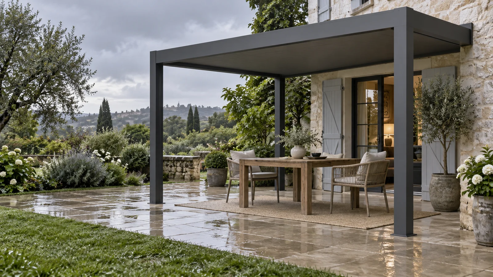 Terrasse humide après la pluie avec table en bois sous une pergola anthracite adossée à une maison en pierre