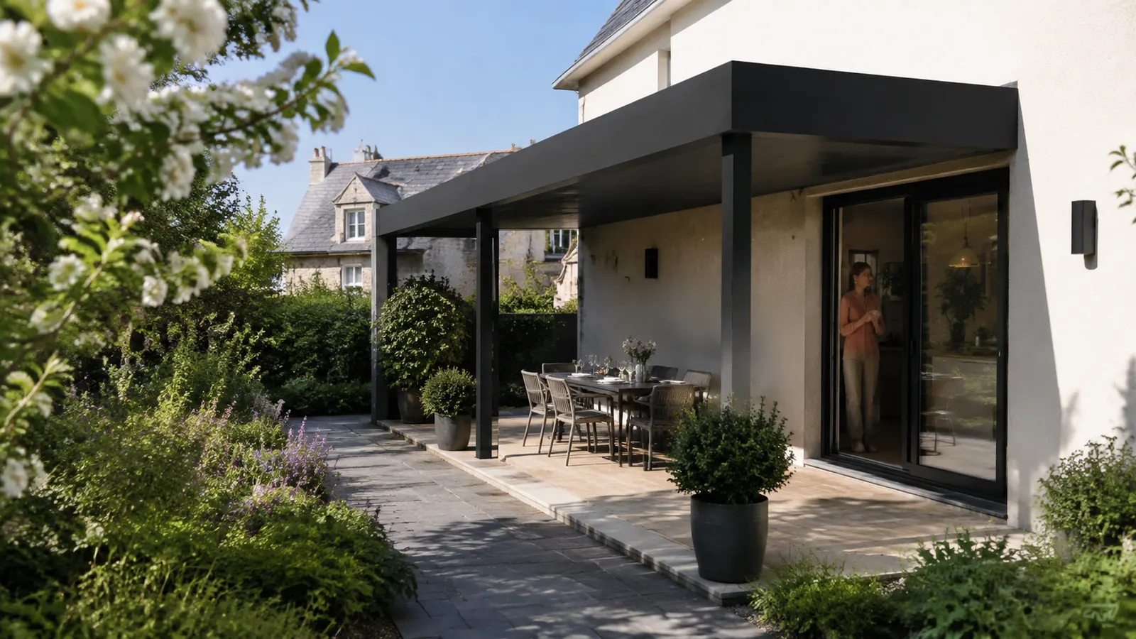 Une baie vitrée ouverte donne sur une terrasse et une pergola anthracite vue depuis le jardin