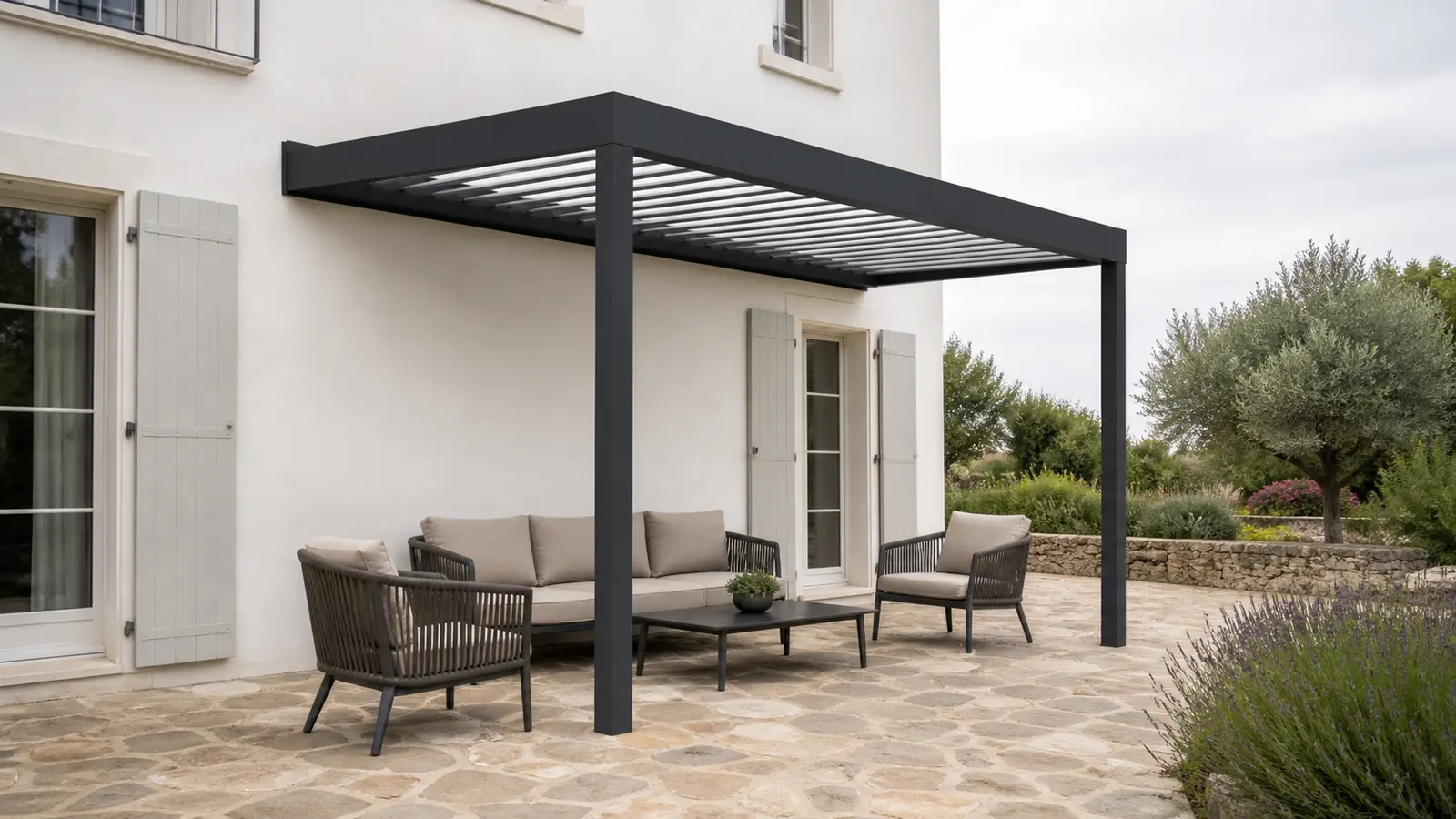 Pergola bioclimatique adossée anthracite avec canapé, deux fauteuils et table basse sur terrasse en pierre