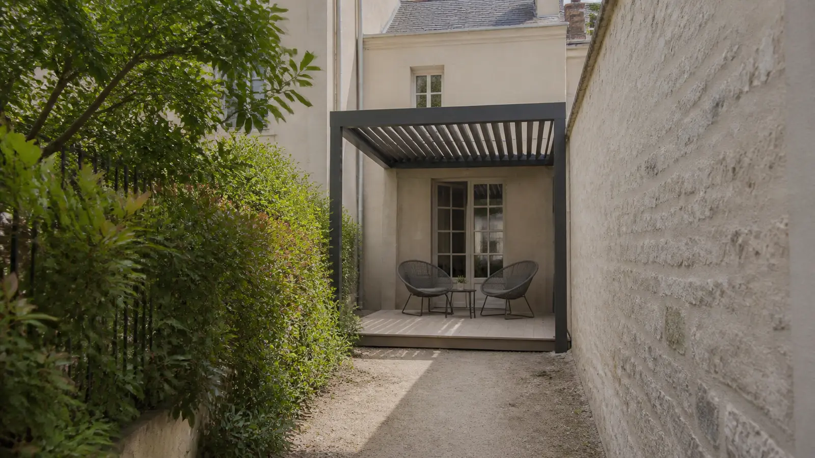 Passage étroit entre un mur et une haie menant à une petite pergola installée dans une cour